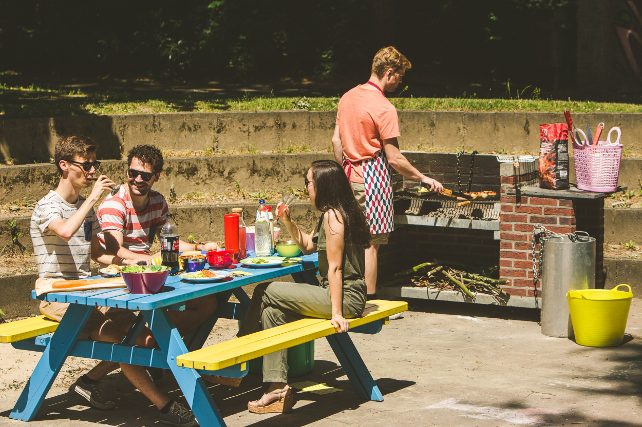 Foto openbare barbecue 1man staat achter de barbecue terwijl de andere 2 aan een picknick tafel zitten te wachten